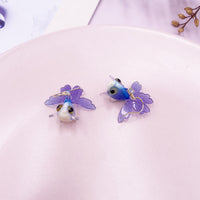 Boucles d’oreilles Poisson Bleu et Violet Transparent, posées sur un fond rose, mettant en valeur leurs nageoires translucides et leur aspect fantaisie.