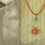 Collier Poisson Orange Pastel avec Étoile de Mer, porté sur un pull torsadé, mettant en valeur un pendentif poisson translucide et des perles marines.