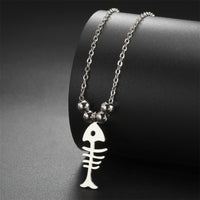 Gros plan sur le Collier Poisson Squelette Argenté à Billes, posé sur un support noir, avec ses perles métalliques et son pendentif minimaliste.