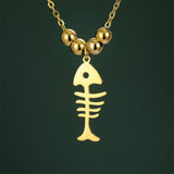 Gros plan sur le pendentif Poisson Squelette Doré à Billes, présenté sur fond vert, soulignant le design minimaliste de l’arête de poisson.