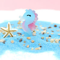 Figurine Hippocampe en résine bleu et rose, posée sur du sable, avec une étoile de mer blanche à proximité.
