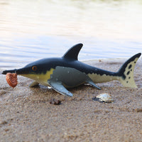 Figurine Mosasaure Préhistorique placée sur le sable au bord de l’eau, avec une coquille d’ammonite dans la gueule, illustrant un décor naturel.
