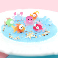 Ensemble de figurines Poisson, crabe et pieuvre en résine, colorées et translucides, disposées sur du sable bleu.
