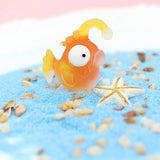 Figurine Poisson Lanterne Orange en résine, posée sur du sable coloré, avec une étoile de mer en arrière-plan.
