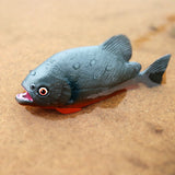 Figurine Poisson Piranha gris et rouge déposée sur un fond sablonneux humide, montrant sa texture détaillée et ses couleurs vives