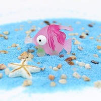 Figurine Poisson Rose en Résine posée sur du sable bleu, avec une étoile de mer blanche en arrière-plan.
