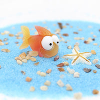 Figurine Poisson Rouge en Résine posée sur du sable coloré, avec une étoile de mer blanche en arrière-plan.
