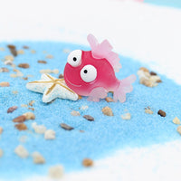 Figurine Poisson Tropique Rouge posée sur du sable bleu, accompagnée d'une étoile de mer blanche.
