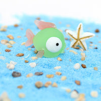 Figurine Poisson Vert en Résine posée sur du sable bleu, avec une étoile de mer blanche en arrière-plan.
