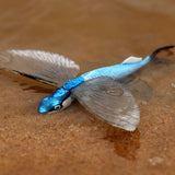 Figurine Poisson Volant – Exocet Bleu posée sur le sable humide, montrant ses nageoires déployées et son dos bleu scintillant.
