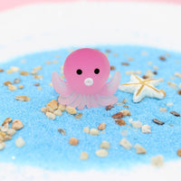 Figurine Poulpe en résine rose, posée sur du sable bleu, avec une étoile de mer en arrière-plan.
